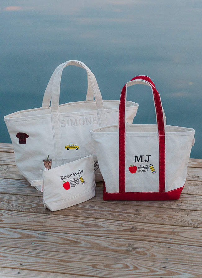 Two white canvas tote bags with red handles and trims, plus a small matching pouch, displayed on a wooden dock with calm water in the background. The larger tote bag features the name 'SIMONS' and small printed illustrations, while the smaller tote features 'MJ' with a red apple and yellow pencil graphic.
